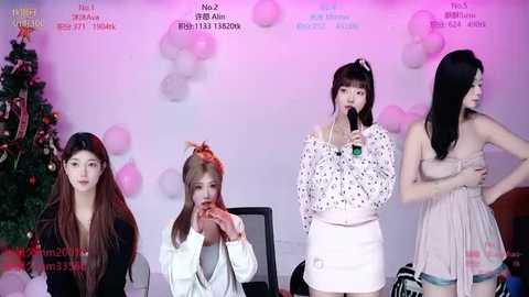 Video of four women on a stage, singing into microphones, wearing festive dresses; Christmas tree and balloons in background.