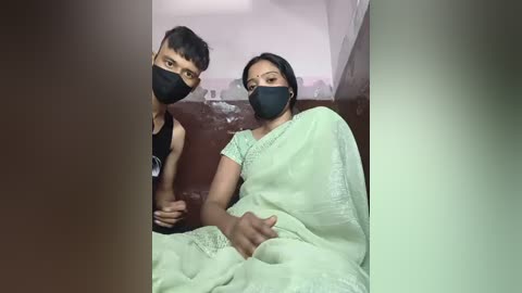 Media: Video of a South Asian couple, wearing black face masks, seated in a dimly lit room with a green curtain. The woman, in a light green sari, has a worried expression; the man, in a black tank top, holds her hand.