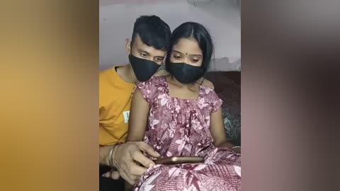 Video of a young Indian couple, both wearing black face masks, sitting close on a sofa. The man in a yellow shirt, holding a phone, gazes at the woman in a pink patterned dress, who looks at him.
