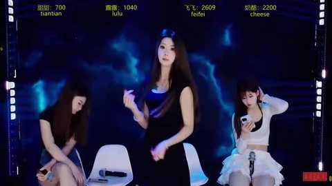 Video of three women in a dimly lit stage setting. The woman in the center, with long black hair, wears a black dress and holds a phone. The others, with short brown hair, sit on white chairs.