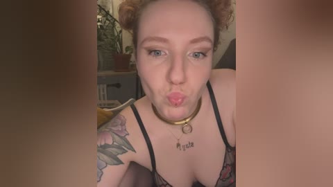 Video of a fair-skinned woman with curly brown hair, wearing a black lace bra, kissing the air. She has a large tattoo of roses on her left shoulder. Background shows a cluttered room with plants.