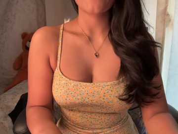 A video of a woman with long, wavy black hair, wearing a floral-patterned tank top, sitting on a bed with beige sheets, and a teddy bear in the background.