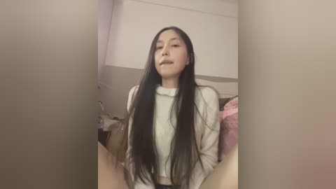 Video of an Asian woman with long black hair, wearing a white turtleneck, seated in a dimly lit room with beige walls and a pink blanket.