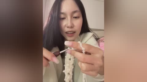 Video of an Asian woman with long black hair applying white nail polish to her nails, wearing a pastel-colored sweater.