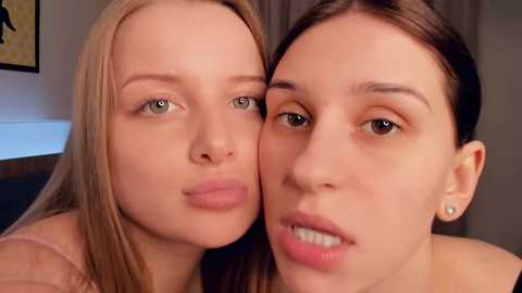 Video of two young women with light skin, green eyes, and straight brown hair. One wears a light pink shirt, while the other has a neutral expression. Background includes a beige wall and a framed painting.