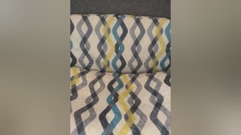 Media: Video of a modern, gray upholstered chair with a cushion featuring a pattern of interlocking, wavy, alternating blue, yellow, and gray lines on a white background. The chair sits against a plain, light beige wall.
