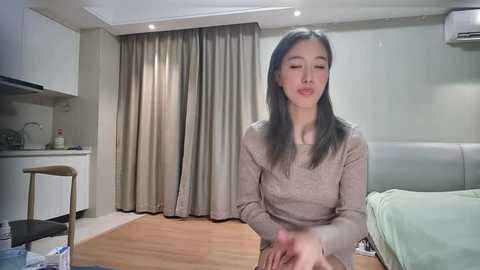 Video of a young East Asian woman with straight, shoulder-length black hair, wearing a beige sweater, seated at a wooden table in a modern, minimalist bedroom with white walls and beige curtains.