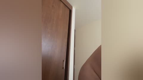 Video of a narrow, beige hallway with a damaged wooden door on the left, a smooth, beige wall on the right, and a partially visible curved staircase leading upward.