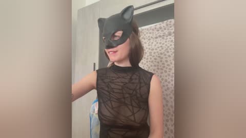 Video of a woman with light skin, wearing a black cat mask and a sheer black mesh top, standing in a bathroom with a leopard print shower curtain.