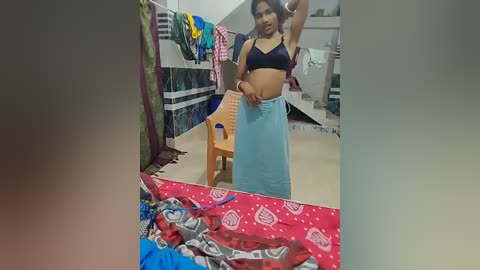 Video of a young South Asian woman in a modest, dark blue bra and light blue sarong, standing in a cluttered, dimly-lit room with clothes hanging on a rack, and a wooden chair.