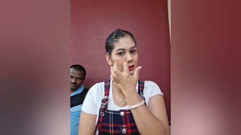 Video of a young woman with dark skin and dark hair styled in a bun, wearing a plaid overalls dress, standing in front of a man in a blue shirt.