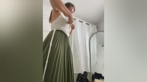 Video of a woman in a white tank top and olive green pleated skirt, adjusting her skirt in a minimalist, softly lit room with white curtains and a large mirror.