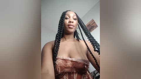 Video of a confident Black woman with medium skin tone and long, braided hair, wearing a strapless, patterned top. Background includes a white wall and a blurred, decorative art piece.