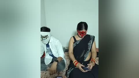 Video of a woman in a black sari and red bindi, sitting on a bed, with a man in a white shirt and blue vest, both with masks, in a dimly lit room.