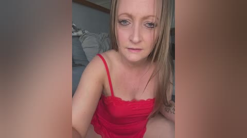 Video of a fair-skinned, blonde woman in a red lace camisole, with a tattoo on her left arm, sitting on a bed with gray sheets, in a casual, indoor setting.