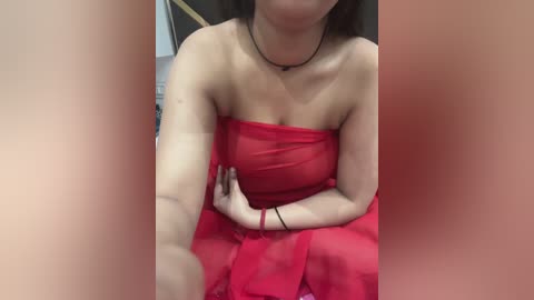 Media: Video of a light-skinned woman with long dark hair, wearing a sheer, strapless red dress, sitting with crossed arms, a black choker, and red bracelet, taken from a close-up angle with blurred background.