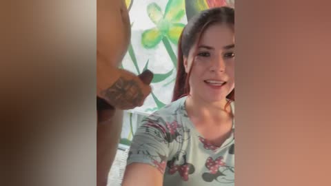 Video of a smiling, dark-haired woman in a floral shirt, partially obscured by two large, erect penises. Background features colorful, abstract art.