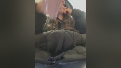 Video: A woman with long brown hair and a black jacket, lying on her side in a train seat, covers her face with her hands, appearing distressed.