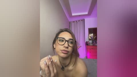 A video of a young woman with light skin, wearing black-framed glasses, and a black necklace, indoors, with purple lighting, looking slightly off-camera.