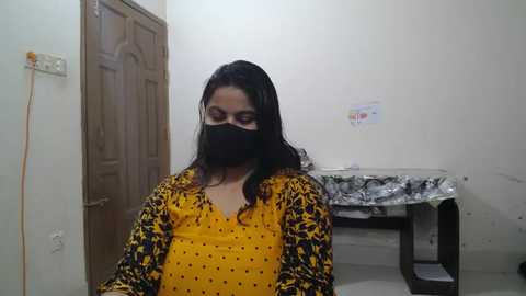 Video of a woman with long dark hair, wearing a yellow top with black polka dots and a black mask, standing in a sparsely furnished room with a wooden door, tiled floor, and a black table.