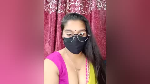 Video of a South Asian woman with medium skin tone, long black hair, wearing a black mask, glasses, and a colorful blouse with a plunging neckline, against a red patterned curtain.