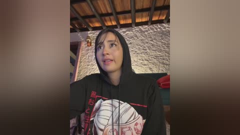 Video of a young woman with long black hair, wearing a black hoodie with a white and red cartoon character, standing indoors with a rustic, wooden-beamed ceiling and a textured, light-colored wall in the background.