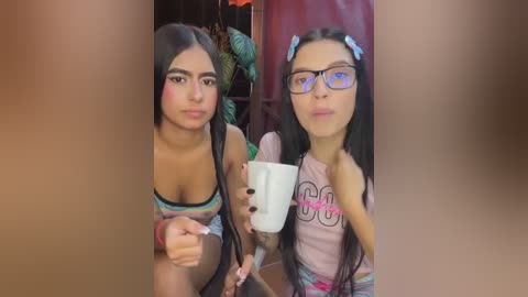 Video of two young women, one with long dark hair and glasses, the other with long black hair and a pink tank top, holding a white cup.