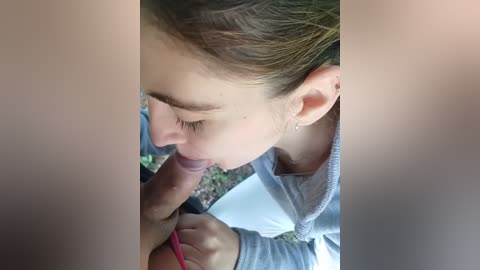 A video captures a close-up of a young woman with light skin and brown hair, kneeling outdoors, performing oral sex on a man. She wears a gray sweater. The background shows grass and a white fence.