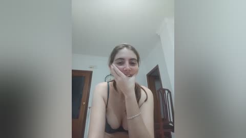 A video of a young, fair-skinned woman with long brown hair, wearing a black bra, leaning against a doorframe, smiling, and touching her face.