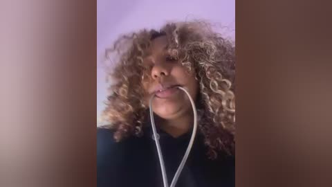 Media: Video of a Black woman with curly, shoulder-length hair, wearing a black top, listening to headphones. The background is a blurred, neutral-colored wall.