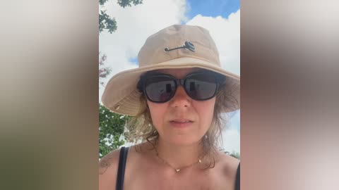 Video of a fair-skinned woman with curly brown hair, wearing a beige bucket hat, black sunglasses, and a black spaghetti strap top, against a backdrop of green foliage and a blue sky with scattered clouds.