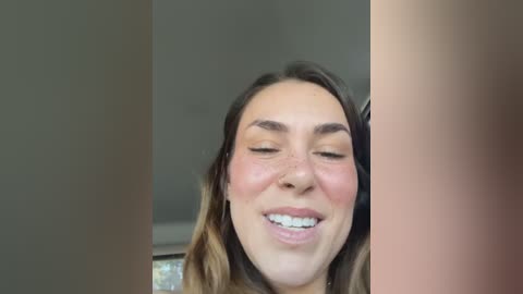 A close-up video of a young woman with fair skin, freckles, and light brown hair, smiling with closed eyes, in a dimly lit room.