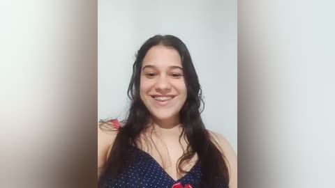 Video of a young woman with long, dark hair, fair skin, and a medium build, wearing a dark blue polka-dot dress, smiling against a plain white wall.
