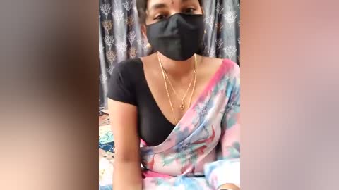 Video of a South Asian woman with medium skin tone, wearing a black mask, black top, and multicolored saree. Background features a patterned curtain.