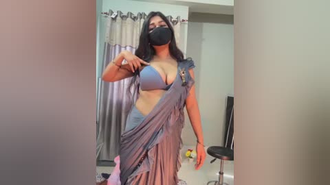 Video of a South Asian woman with medium skin tone, black hair, wearing a blue bra, gray saree, and black mask, standing in a modern bedroom with beige curtains and a black office chair.