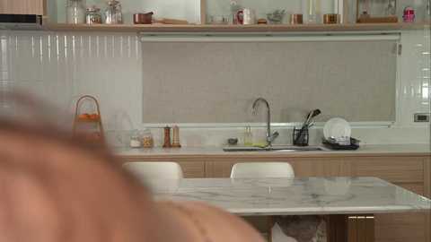 A video of a modern kitchen with a white marble countertop, stainless steel appliances, and wooden cabinets. A person with short, light brown hair is partially visible in the foreground.