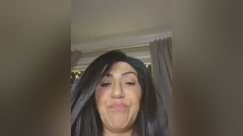 Video of a smiling, dark-haired woman with a slightly distorted face, wearing a nose ring. She stands indoors near a window with light-colored curtains, creating a soft, diffused background.