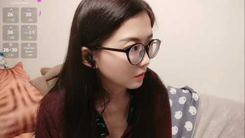 Video of a young woman with long black hair, wearing glasses and a green necklace, sitting on a couch. Background shows a gray pillow with white polka dots and a beige blanket.