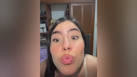Video of a young woman with light skin, dark hair, and full lips puckered for a kiss. She wears a white tank top in a cluttered room with wooden cabinets and a bookshelf.