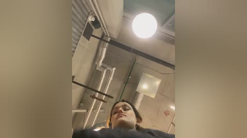 Video of a young woman with light skin and dark hair, standing in a modern, industrial bathroom with metallic fixtures and a large, circular ceiling light. She looks down at a sink and mirror.