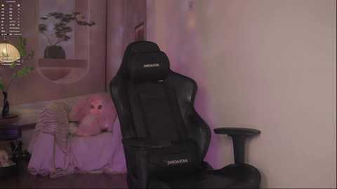 Video of a black gaming chair with a logo, in a cozy, dimly-lit room with a plush pink couch, a green plant, and a lamp.