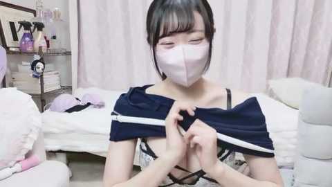 A young East Asian woman with short black hair and a white surgical mask, dressed in a black lace bra, is removing a blue top in a cluttered bedroom with pink curtains and various personal items.