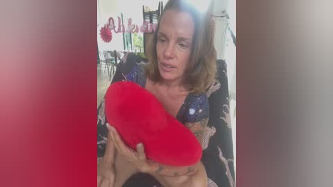 A video of a middle-aged woman with shoulder-length brown hair, wearing a black floral-patterned top, holding a red heart-shaped pillow close to her chest. The background features a blurred room with red and white decorations.