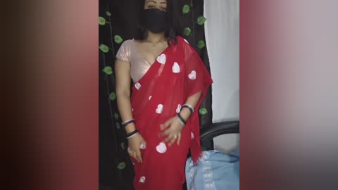 Video of a dark-skinned woman with long black hair, wearing a red saree with white hearts, black mask, and black wristbands, standing in a dimly lit room with green garlands.