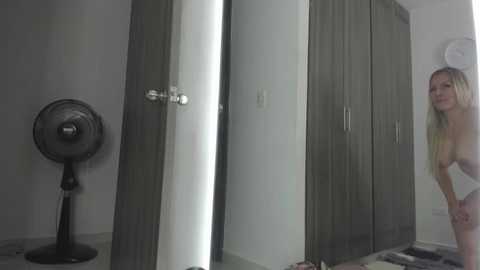A video of a nude blonde woman with small breasts standing next to a closed door in a dimly lit room with wooden cabinets and a black oscillating fan.