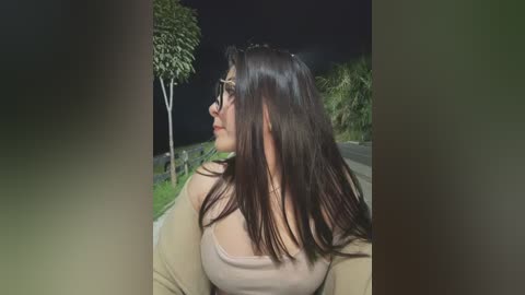 Video of a young woman with long brown hair, wearing glasses, a beige top, and a beige cardigan, gazing at a tree-lined road at night.