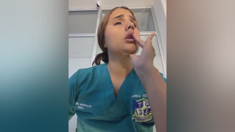 Video of a young woman with light brown skin, medium build, wearing teal scrubs with \"Lana M. Aguilar, RN\" embroidered on the chest, biting her finger, in a clinical setting with white walls and a door.