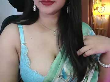 Media: Video of a light-skinned woman with long black hair, wearing a light blue lace bra and a green sari with floral patterns. She has red lipstick and pearl earrings, sitting in a chair with a warm, lit room in the background.