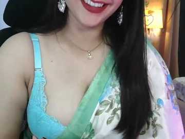Media: Video of a smiling, fair-skinned woman with long black hair, wearing a turquoise lace bra, green floral sari, silver earrings, and a gold necklace, set indoors with a warm, soft-focus background.