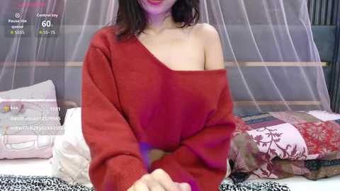 Video of a fair-skinned woman with dark hair, wearing an off-shoulder red sweater, sitting on a bed with floral-patterned bedding, surrounded by pillows and a sheer canopy.